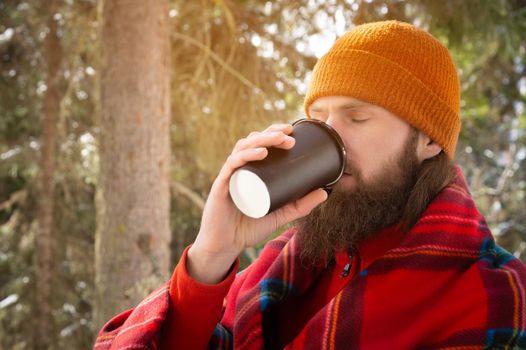 portrait of a caucasian bearded man in a hat and wrapped in a plaid, drinking a drink from a paper cup, against the backdrop of a coniferous forest