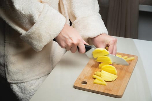 young caucasian woman in a bathrobe stands in the kitchen and cuts fresh potatoes with a knife, on a wooden cutting board