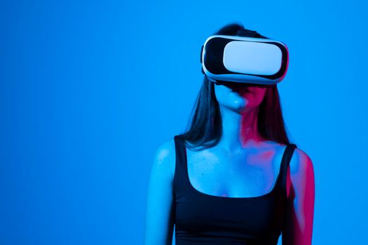 Concept of modern gadgets and technologies. Future technology concept. Portrait of young woman in VR-glasses in neon light on blue background. Virtual reality gaming