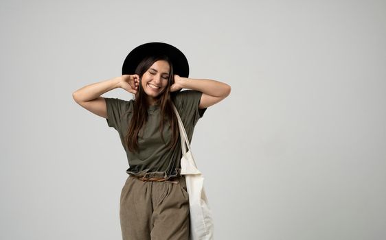 Zero waste, plastic free concept. Happy attractive woman in green t-shirt and black hat holding cotton shopper bag with vegetables, products in white room. Eco friendly shopping bags