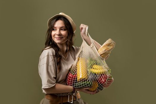 Smiling young woman in light summer clothes with a mesh eco bag full of vegetables, greens on a green studio background. Sustainable lifestyle. Eco friendly concept. Zero waste