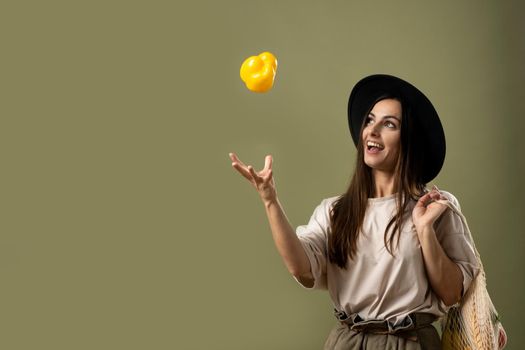 Zero waste concept. Eco friendly happy smiling woman in beige t-shirt and black hat throw up a yellow pepper in a air by one hand and holding reusable cotton eco bags full of groceries on a shoulder