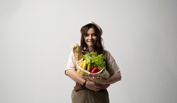 Eco friendly positive young woman in beige oversize t-shirt holding reusable mesh cotton eco bags for shopping with organic groceries on white background