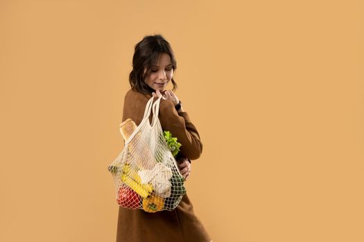 Back view of happy smiling young woman in brown dress holding reusable string bag with organic fruits and vegetables over orange background. Sustainability, eco living and people concept
