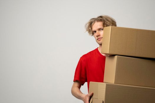 Young delivery man in a red uniform holding a stack of cardboard boxes. Courier delivering postal packages, parcels over white studio background