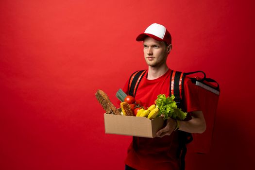 Delivery male employee in a red uniform holds paper cardboard package with organic groceries, fruits on a red background. Products delivery from shop or restaurant to home. Courier service