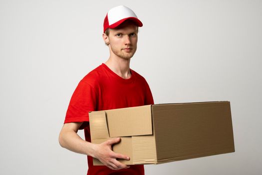 Delivery man in a red uniform and a cap holding cardboard box delivering postal package and ready to deliver it to a client over white studio background