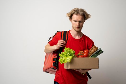 Food delivery concept. Delivery service from groceries market to home. Courier in red t-shirt and thermal bag backpack on a shoulder holds a paper box with organic foods, products