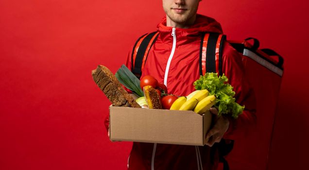 Male courier in a uniform holding a grocery box with organic food and bring it to a customer. Express food order. Food delivery service