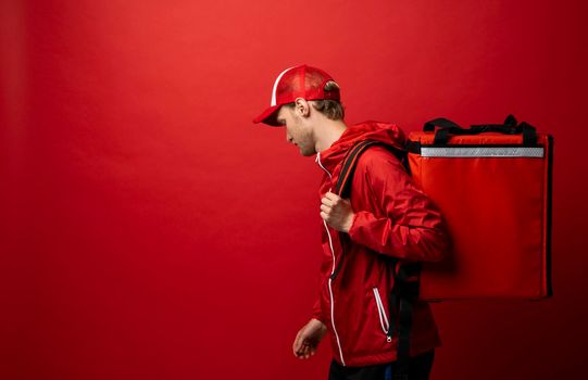 Side view of young professional courier in a red uniform and with a thermal bag on a shoulder on color red background, space for text. Food delivery service