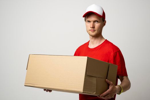 Delivery service. Young courier in a red uniform holding cardboard box. Happy young delivery man in cap and red t-shirt standing with parcel isolated on white background