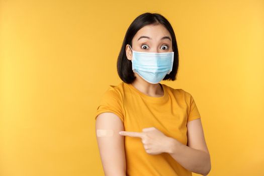 Vaccination from covid and health concept. Excited asian woman in medical face mask, pointing finger at shoulder with band aid, standing over yellow background.