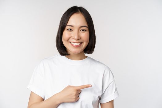 Happy beautiful asian girl showing advertisement on empty copy space, pointing finger right and smiling, demonstrating banner or company logo, white background.