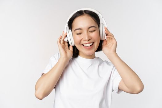 Modern asian girl dancing, listening music with headphones, smiling happy, standing in tshirt over white background.