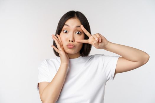 Cute asian girl posing with kawaii v-sign, peace gesture near face, standing in tshirt over white background. Copy space