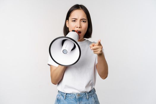 Angry asian woman with megaphone, scolding, accusing someone, protesting with speakerphone on protest, standing over white background.