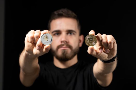 Young bearded man holding virtual currency ethereum coin in one hand and bitcoin coin in another on a black background. Cryptocurrency