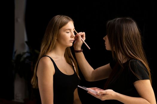 Makeup artist applies eye shadow on a beautiful woman face. Hand of visagiste, painting cosmetics of young beauty model girl. Make up in process