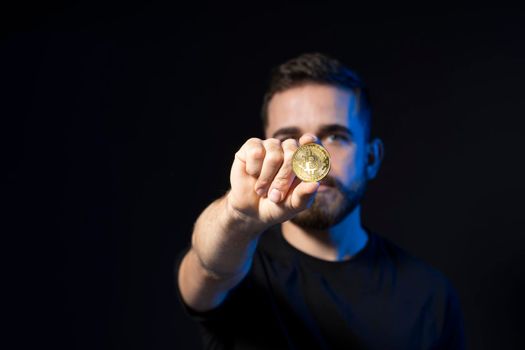 Young handsome bearded successful businessman in black t-shirt shows golden bitcoin on camera isolated on black background