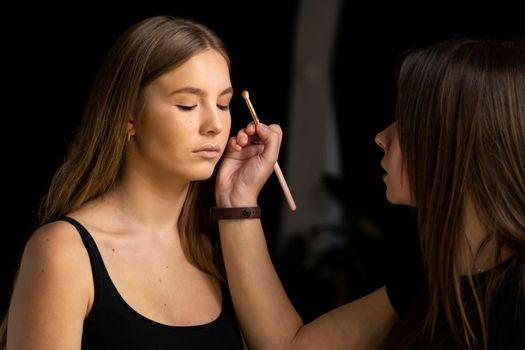 Make-up artist applying eyeshadow in the corner of model eyes and holding a shell with eyeshadow in a black studio. Professional make up