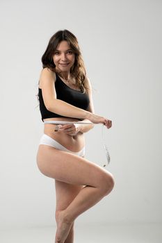 Portrait of a satisfied happy attractive sexy brunette fitness woman in a underwear holding measuring tape around her waist isolated over white background