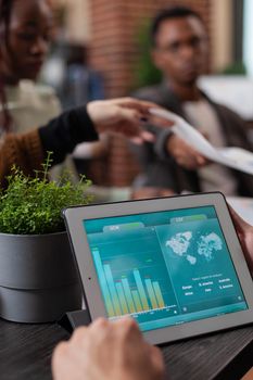 Closeup of entrepreneur holding tablet computer with marketing statistics on screen. Multiethnic businesspeople working at marketing project sharing paper with company turnover during business meeting