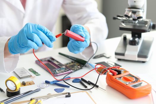 A man with a voltmeter and a red battery in his hands, close-up. Tools and parts for building a device