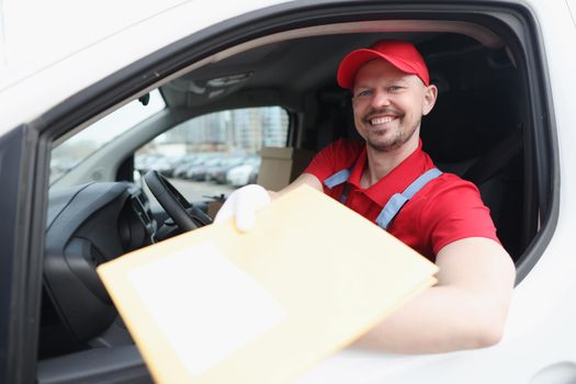 A courier in a red uniform sitting in a car holds out a package, close-up. Delivery of cargo, distribution, representative