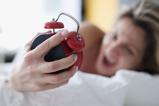 Screaming woman in bed with a red alarm clock in her hand, close-up, blurry. Oversleep for work, morning stress