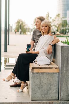 An adult mature happy couple in love sitting on bench outdoors in city street park. A blonde caucasian man and woman spend time together and drinking coffee. Senior wife and husband walking outside