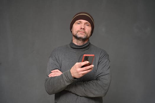 Caucasian adult man in a hat with a phone looks at the camera on a gray background