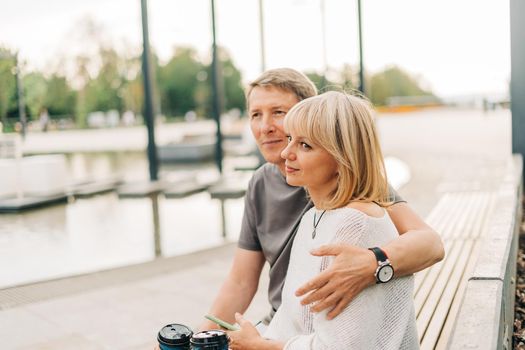 An adult mature happy couple in love sitting on bench outdoors in city street park. A blonde caucasian man and woman spend time together and drinking coffee. Senior wife and husband walking outside