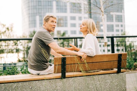 An adult mature happy romantic couple in love sitting on bench outdoors in city street park. A blonde caucasian man and woman spend time together. Senior wife and husband walking outside