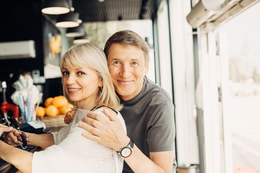 An adult mature happy couple in love in a street cafe order coffee. A blonde caucasian man and woman spend time together. Senior wife and husband walking outdoors