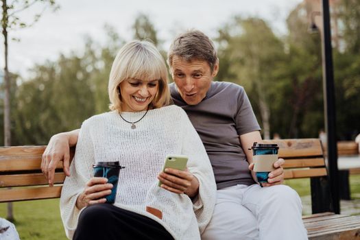 Laughing adult mature couple in love sitting on bench with phone outdoors in park. A blonde caucasian man and woman spend time together and drinking coffee. Senior wife and husband walking outside