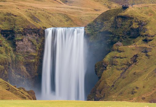 Silk water of Skogafoss waterfall in Iceland from the distance with hikers on top viewpoint