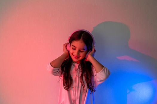 Beautiful happy little girl in pink headphones with loose dark hair listening to music in neon pink blue light holding headphones with her hands