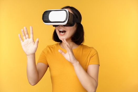 Girl in VR. Beautiful young asian woman, using virtual reality glasses and playing, chatting virtually, standing over yellow background.