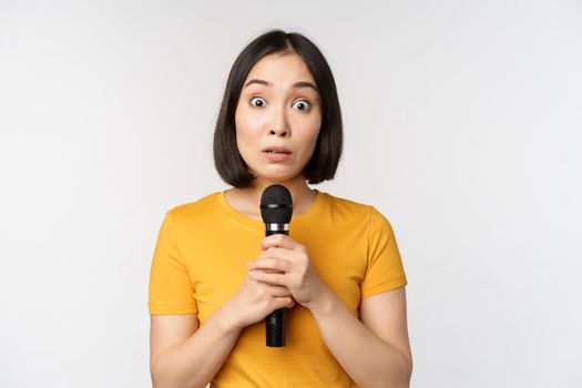 Modest asian girl holding microphone, scared talking in public, standing against white background. Copy space