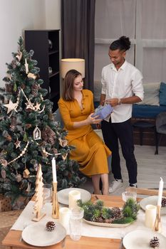 A young man in a white shirt and black pants, gives a lilac box with a gift to a young beautiful slender woman in a mustard dress, in a modern living room, next to a Christmas tree and a table set
