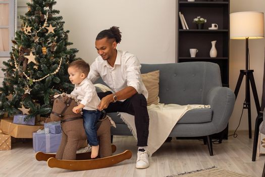 A young stylish father sits on a gray sofa, rolls his little son on a brown rocking horse, at home, in the evening. They smile.