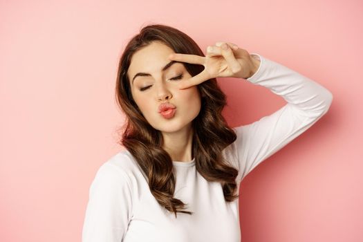 Close up portrait of coquettish young woman smiling, showing peace, v-sign gesture and posing happy, standing against pink background.