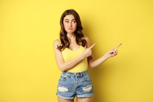 Skeptical woman pointing, looking right with doubtful, uncertain face expression, hesitating, standing over yellow background.
