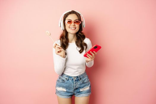 Stylish brunette woman dancing in headphones with mobile phone, listening music in earphones, licking lolipop, standing over pink background.