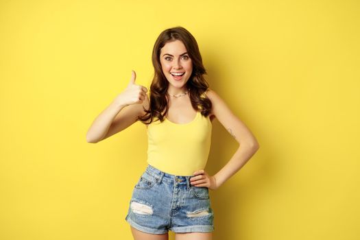 Portrait of stylish modern girl, feminine woman showing thumbs up, recommending gesture, like or approve, praise, standing over yellow background.