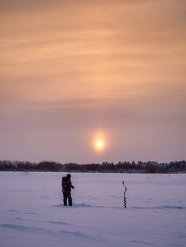 A young man fishes from a hole in the ice at dawn. winter fishing