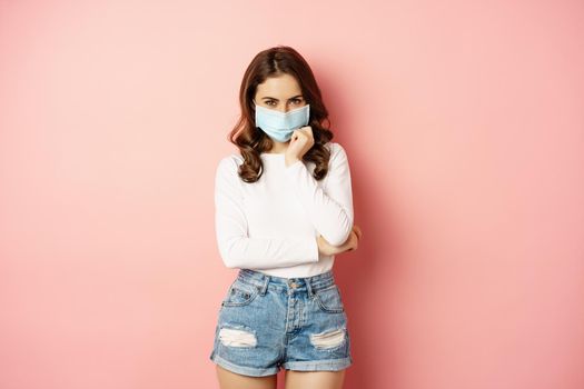 Portrait of stylish modern girl in medical face mask, looking coquettish and smiling at camera, standing over pink background. Pandemic and covid concept.