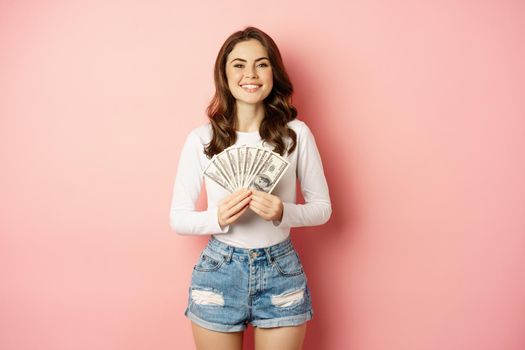 Loans and microcredit. Smiling beautiful girl showing money, cash in hands and looking enthusiastic, standing over pink background.