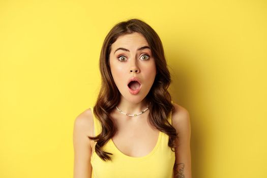 Close up portrait of stylish brunette girl looking amazed, wow face, showing excitement and surprise, impressed by something, standing over yellow background.