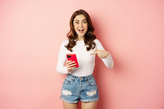 Happy brunette girl showing announcement, pointing finger at mobile phone, showing online promo, app news, standing against pink background.